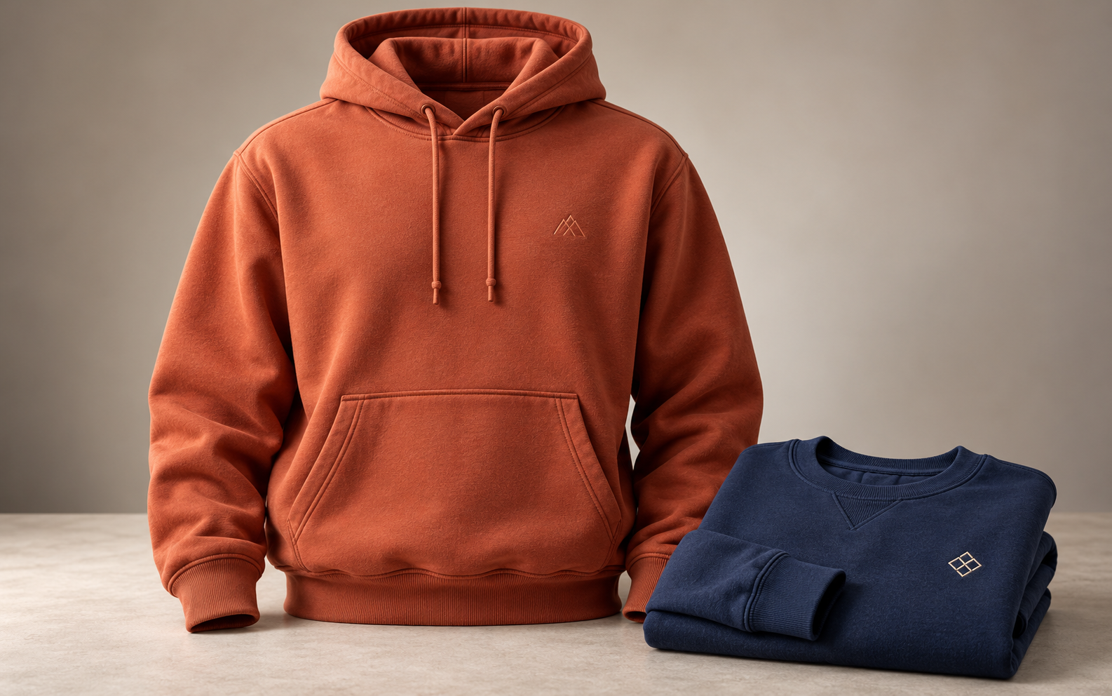 Sudadera terracota con capucha y bolsillo cangurera junto a una sudadera azul marino doblada